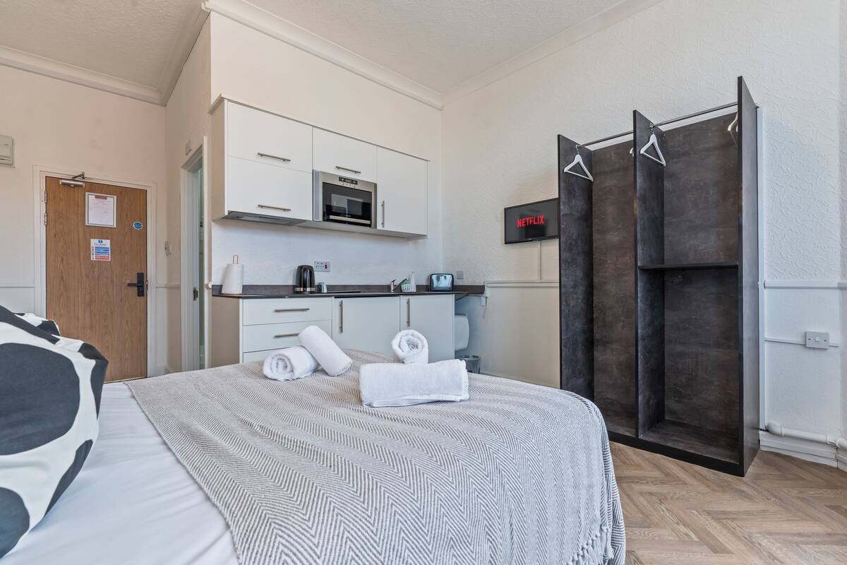 A hotel room with a bed, a kitchenette, a shower, and a TV displaying the Netflix logo.