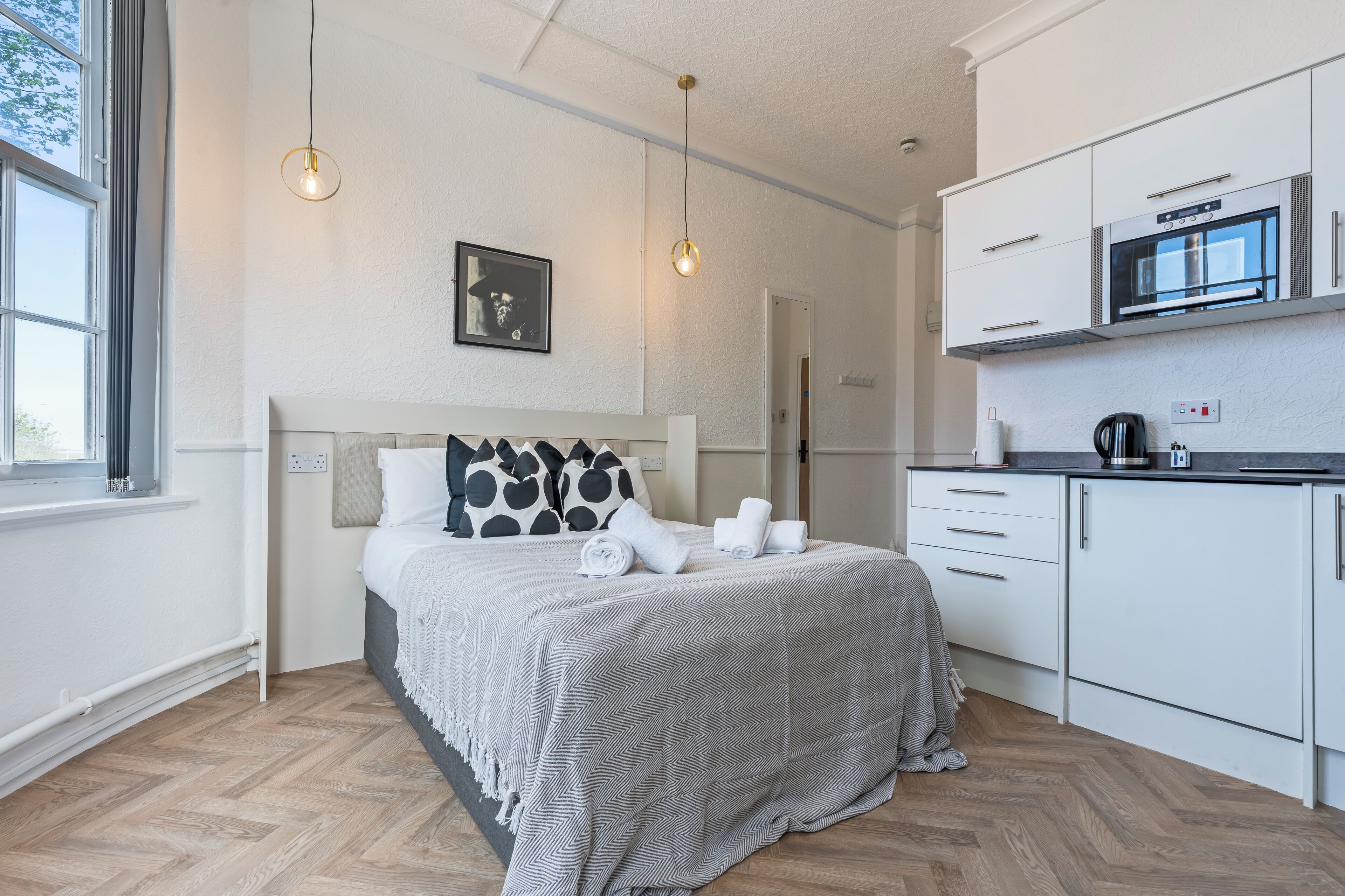 A modern hotel room with a bed, a kitchenette, and a large window.