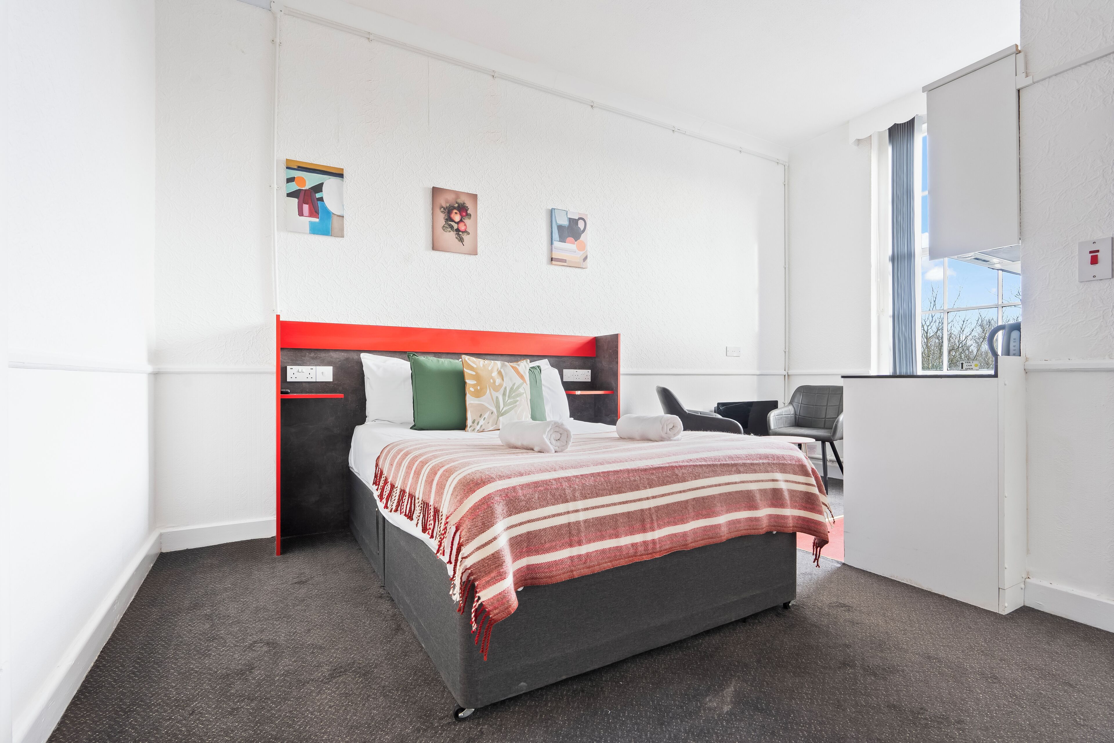 A modern bedroom with a grey bed, red headboard, and a striped blanket. There are two pillows, a green pillow, and a white pillow. The room has white walls, a window with blue curtains, and a small desk with a chair.