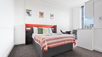 A modern bedroom with a grey bed, red headboard, and a striped blanket. There are two pillows, a green pillow, and a white pillow. The room has white walls, a window with blue curtains, and a small desk with a chair.