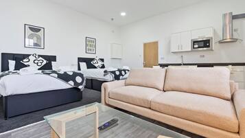 A modern hotel room with two beds, a sofa, a glass coffee table, a microwave, and a cabinet.
