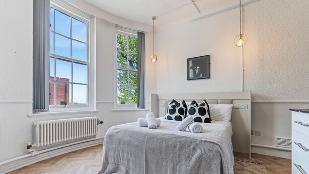 A bedroom with a large window, a radiator, a bed with a patterned bedspread, two bedside lamps, and a wall-mounted picture. - Compact Studios in Dudley (Dudley)