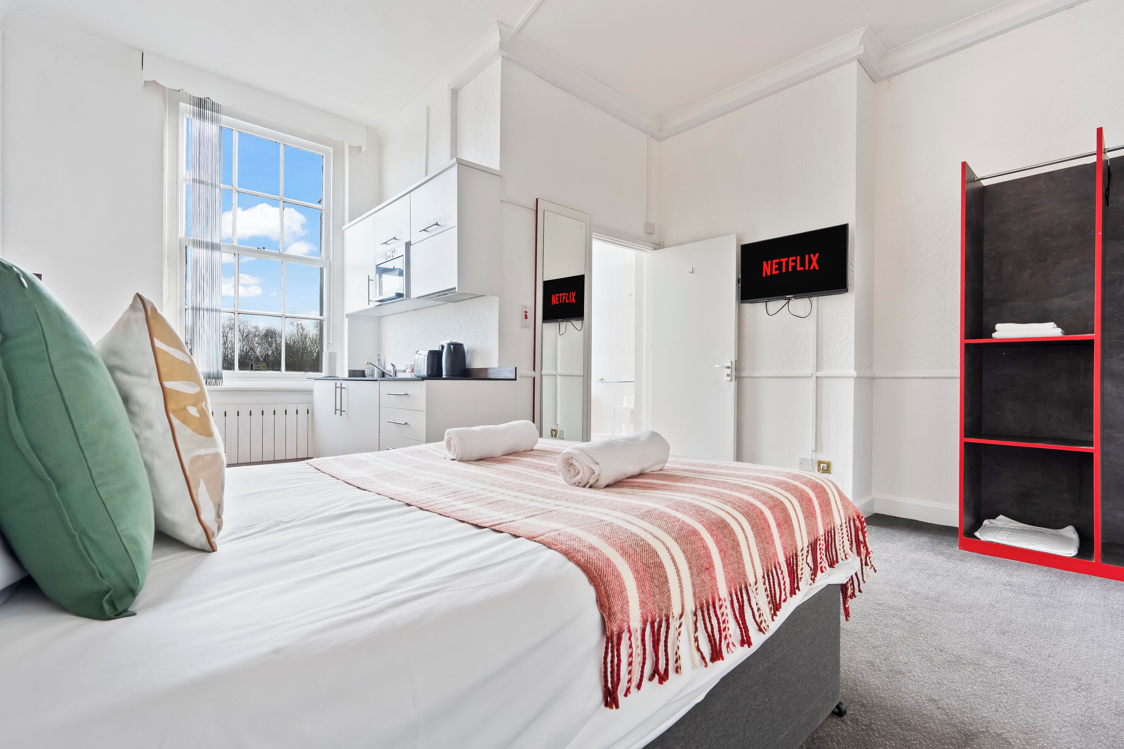 Una habitación de hotel moderna con una cama, un armario, una kitchenette y una pantalla de televisor mostrando 'NETFLIX'.