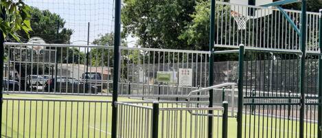 Una cancha deportiva con un aro de baloncesto, rodeada de vallas metálicas y árboles.