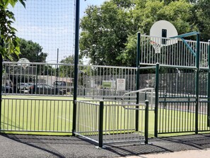 Ein Sportplatz mit Basketballkorb, umgeben von Metallzaun und Bäumen.