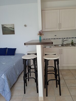 Eine kompakte Küchenzeile mit Essbereich, inklusive einem Bett mit blau-weiß gestreifter Bettdecke, einem kleinen Tisch mit Hocker und einem wandmontierten Spülbecken.