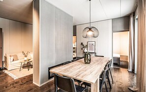 A modern interior with a wooden dining table, black chairs, a hanging light fixture, and a view into a living area with a sofa and a coffee table.