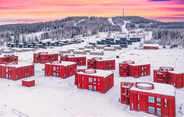 A snowy landscape with red buildings, a ski slope, and a forested hill in the background.