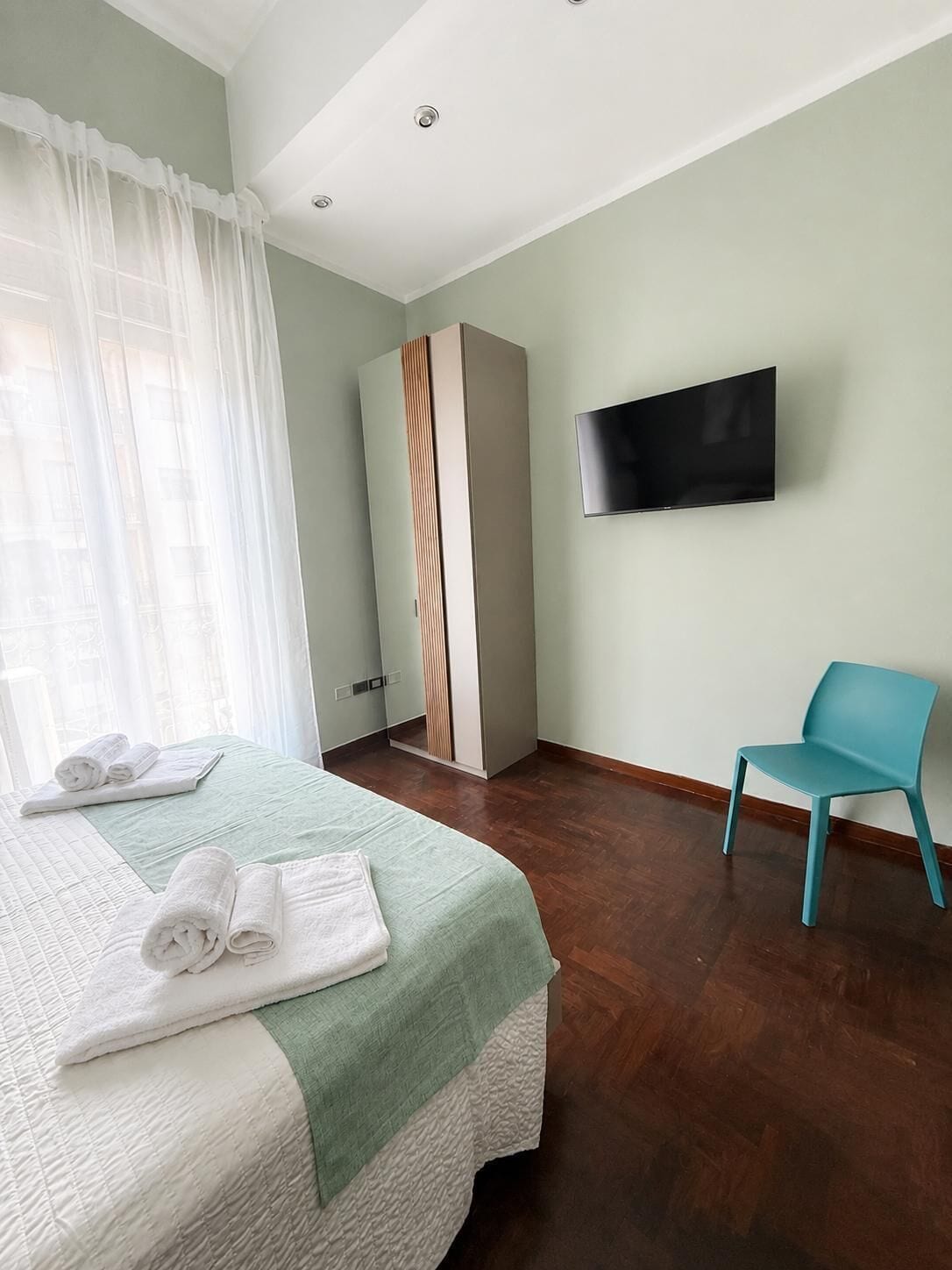A hotel room with a bed, a TV mounted on the wall, a blue chair, and a wardrobe.