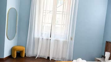 A hotel room with a bed, a window with sheer curtains, a wooden floor, and a small stool.