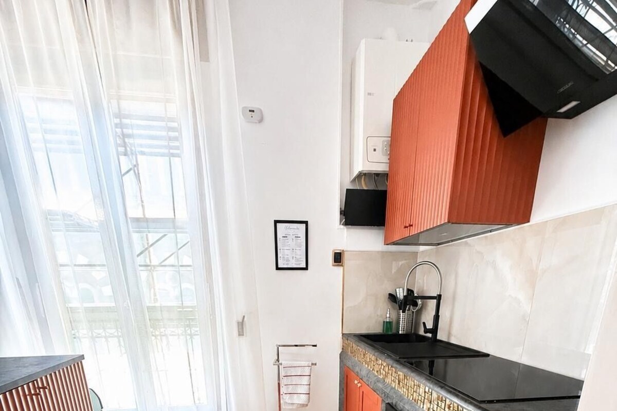 Una cocina moderna con horno empotrado, pileta y estufa. Posee piso de madera y un ventanal con cortinas traslúcidas.