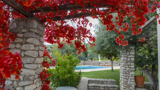 Une pergola en pierre ornée de fleurs rouges, une piscine et une terrasse avec une table et des chaises.