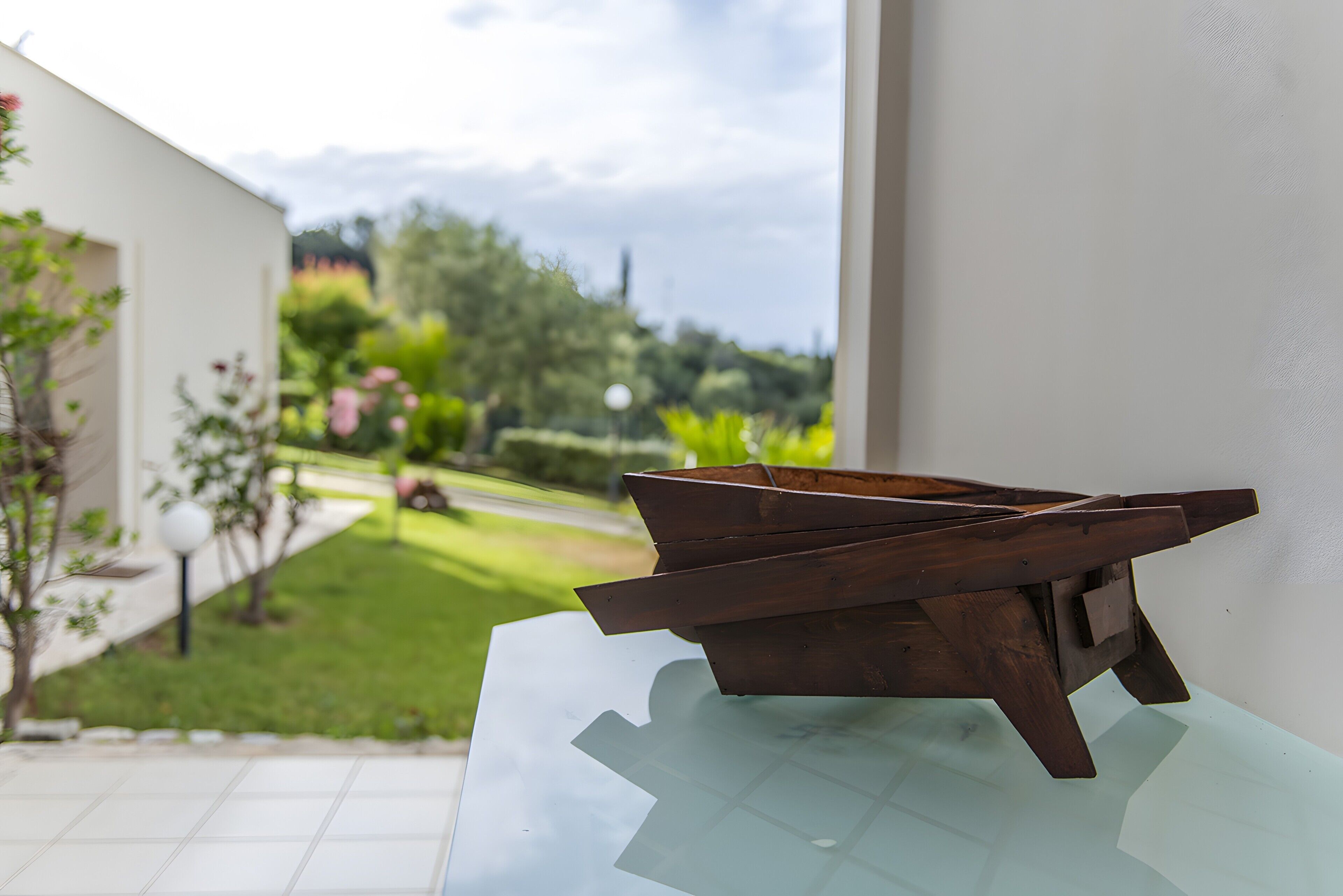A wooden table with a glass top, a white wall, and a view of a garden with a pathway and trees.