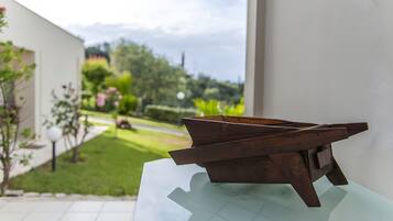A wooden table with a glass top, a white wall, and a view of a garden with a pathway and trees.