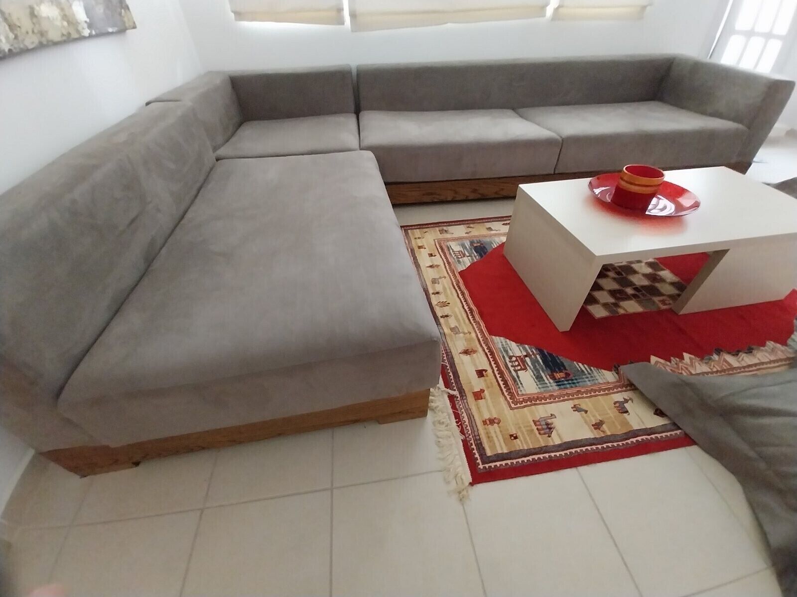 Ein Wohnzimmer mit einem grauen Ecksofa, einem weißen Couchtisch und einem roten und braunen Teppich sowie einem Fenster mit Jalousien.