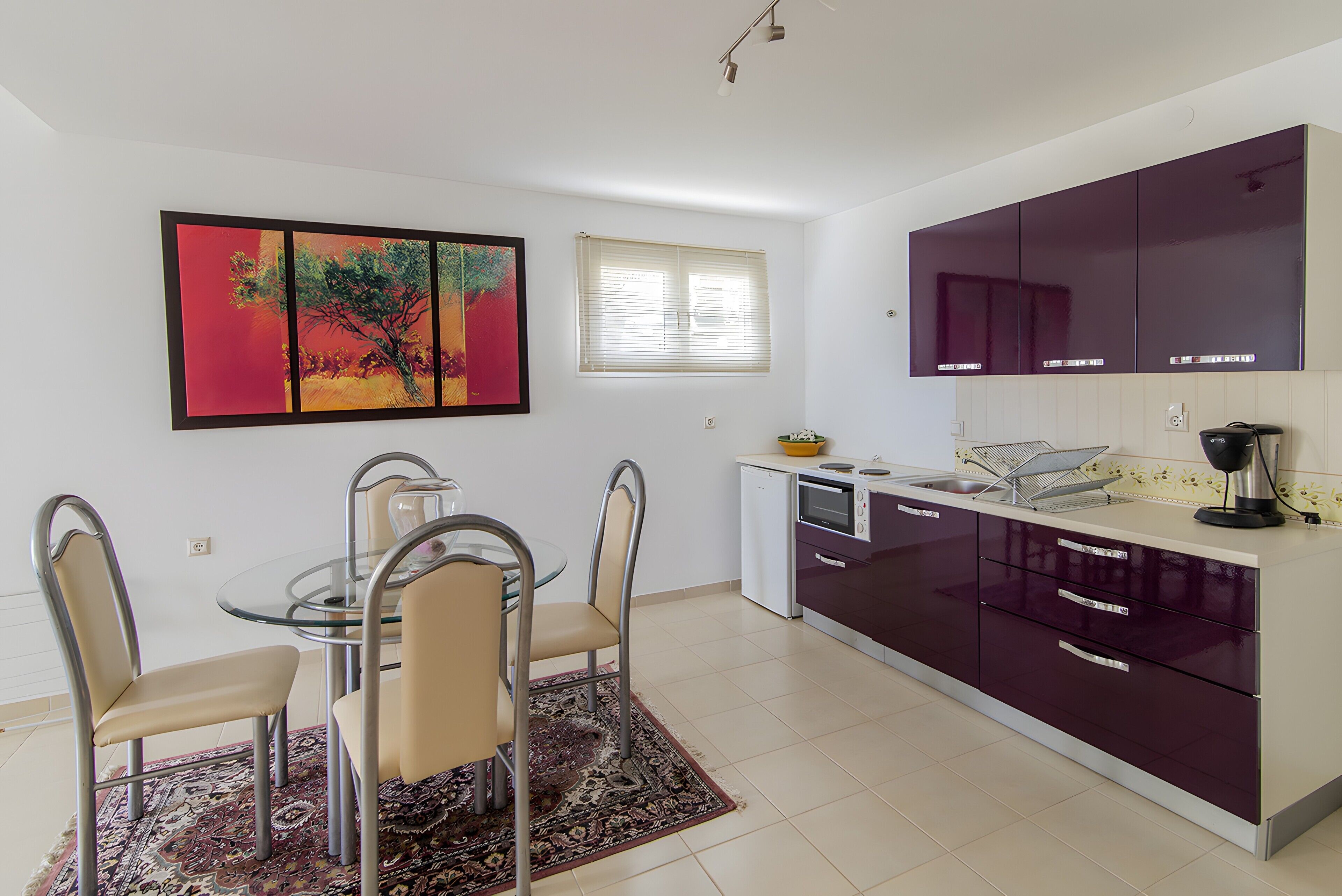 Una cucina moderna con zona pranzo, con un tavolo in vetro, sedie metalliche, un quadro di grandi dimensioni e una finestra con persiane.
