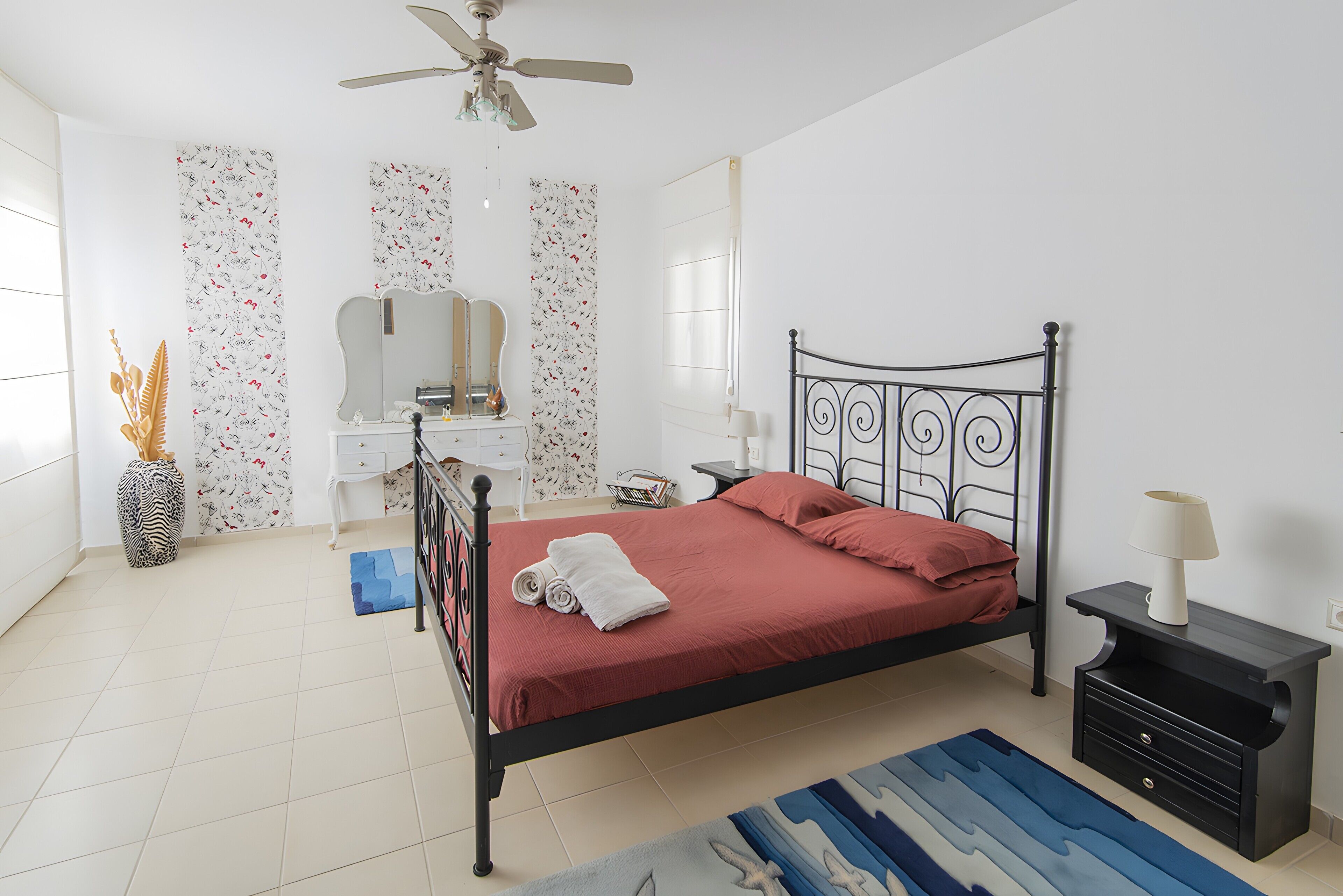 A bedroom with a bed, bedside table, nightstand, mirror, dresser, and a ceiling fan.