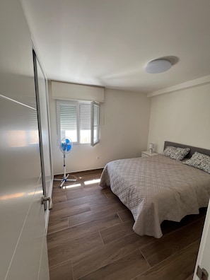 A bedroom with a bed, bedside table, lamp, window with blinds, and a standing fan.