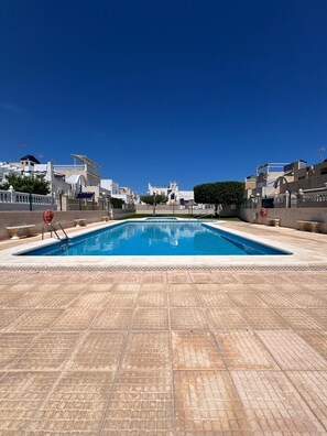 Une piscine rectangulaire bordée d’une terrasse carrelée, entourée d’une clôture, et un ciel bleu dégagé au-dessus.