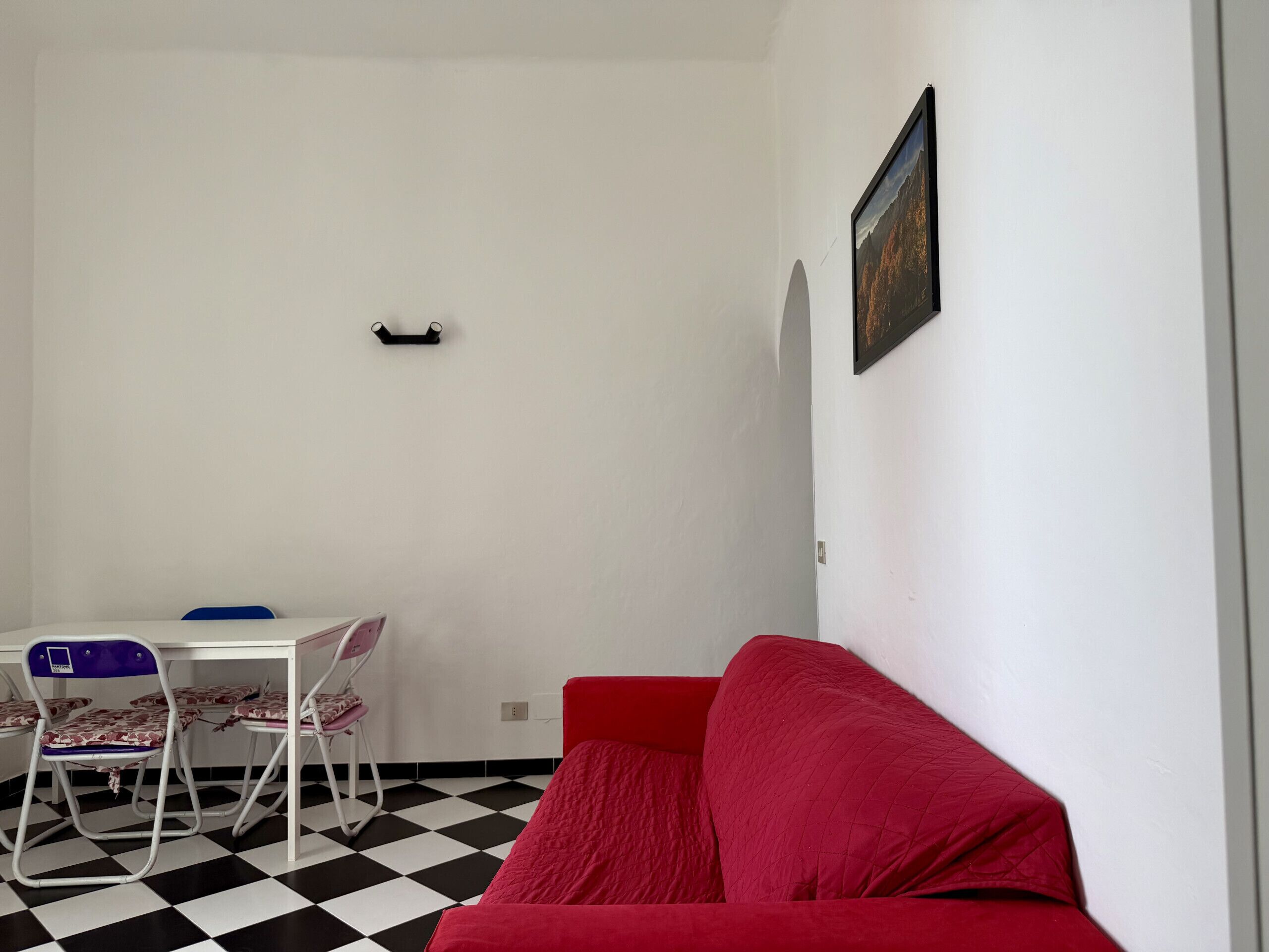 Een kamer met een rode bank, een witte tafel met stoelen, een zwart-wit geruit vloertje en een lijst met een foto aan de muur.