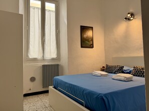 Ein Schlafzimmer mit Bett, Heizkörper, einem Vorhangfenster, einer Wandleuchte und einem gerahmten Bild.