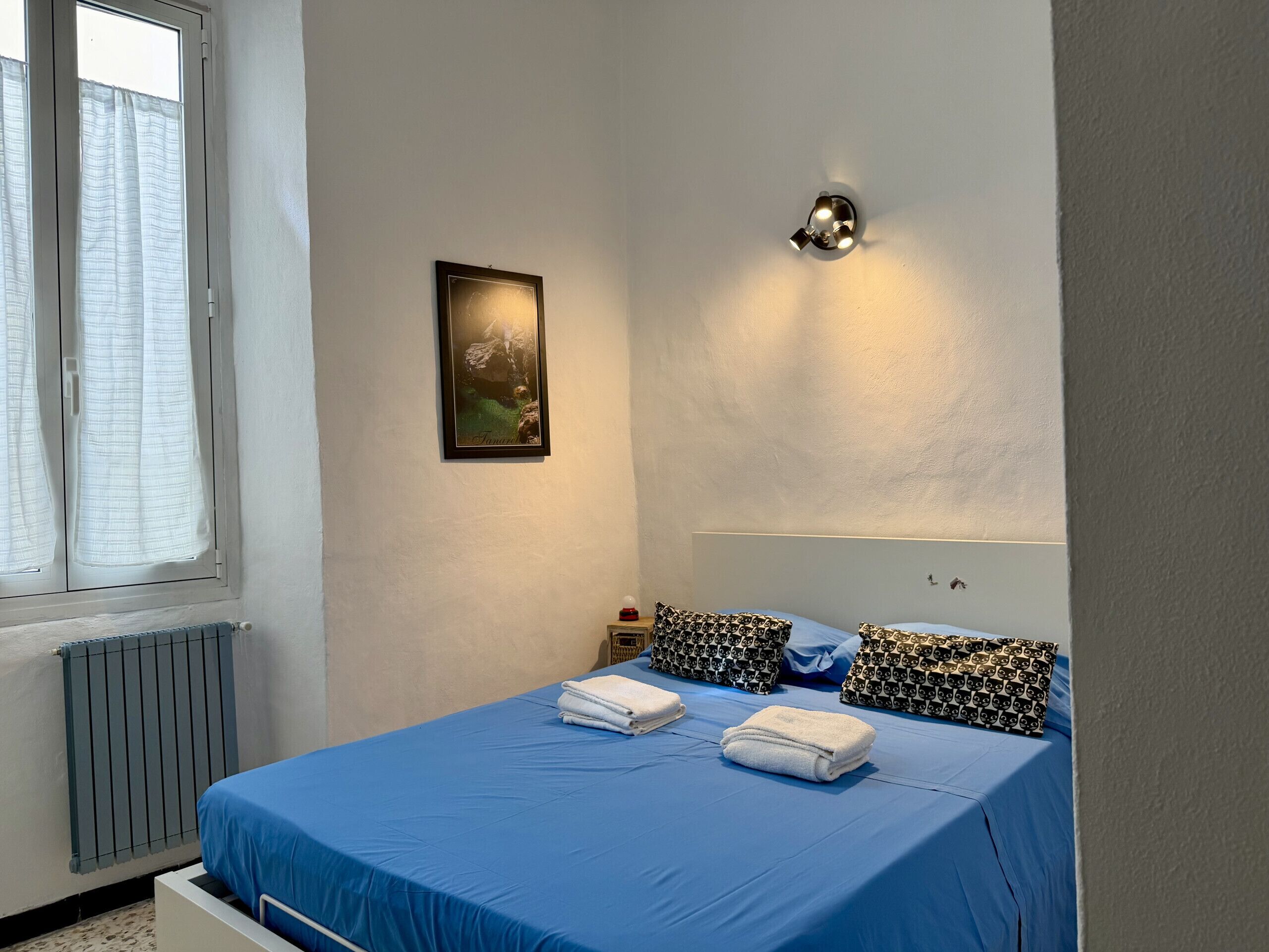 A bedroom with a bed, a radiator, a window with curtains, a wall-mounted light, and a framed picture.
