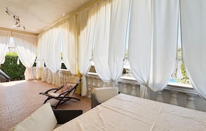 Ein Zimmer mit Balkon, weißen Vorhängen, einem Korbstuhl und einem Tisch.