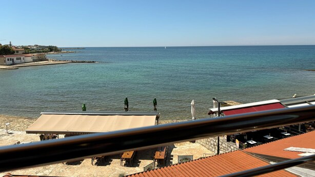 Vista al mar con una terraza de restaurante con vistas al océano, un velero a lo lejos y cielo despejado.