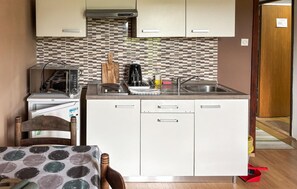 Een kleine keuken met een magnetron, broodrooster en koffiezetapparaat, een spoelbak, en een eethoek met een tafel en stoel.