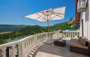 Ein Balkon mit weißer Brüstung, einem Korbsessel, einem Tisch mit einem Getränk und einem großen Sonnenschirm.
