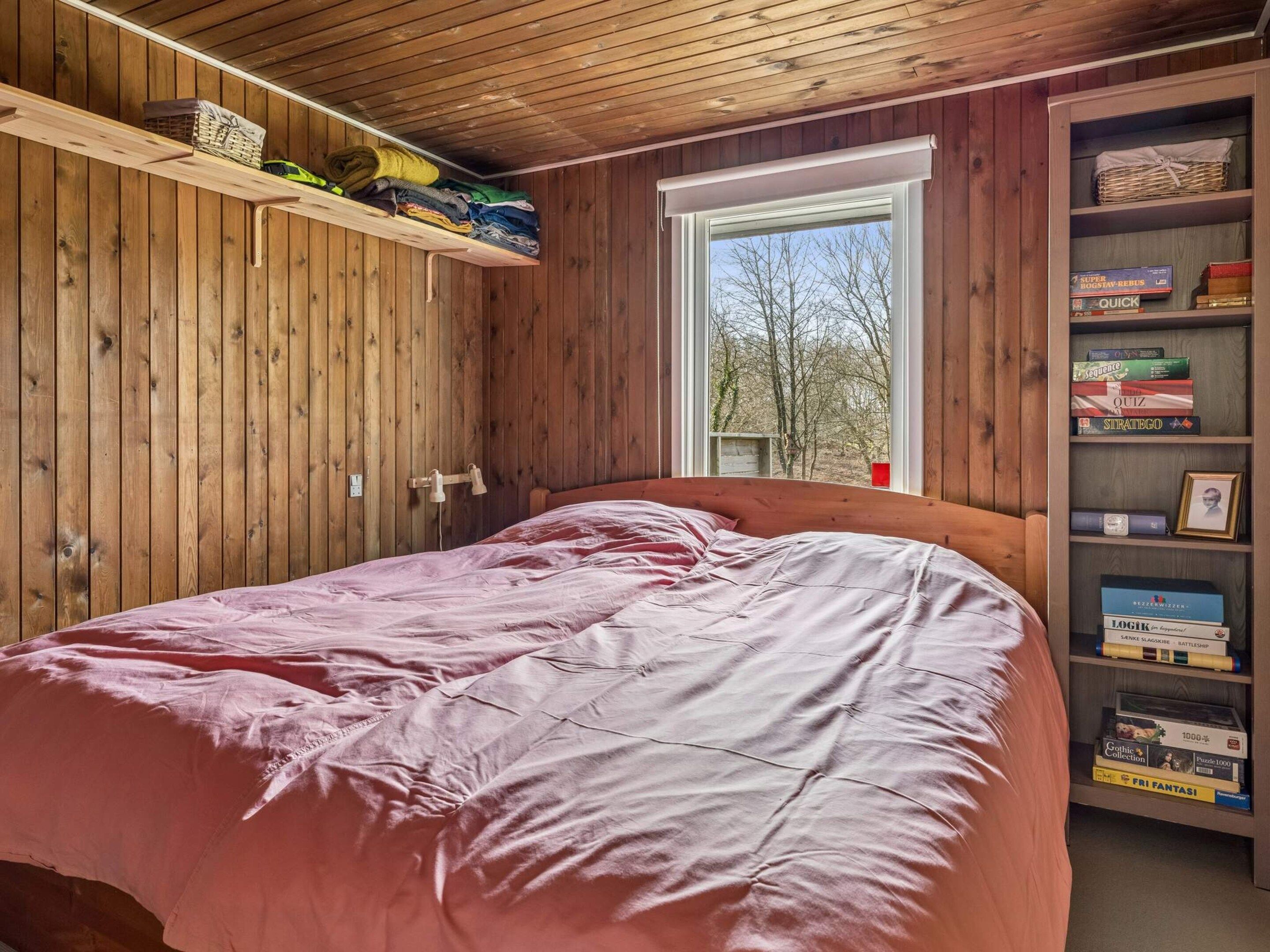 Un dormitorio con techo de madera, una ventana con vistas a árboles, una cama con ropa de cama rosa y una estantería con varios libros y objetos.