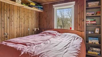 Un dormitorio con techo de madera, una ventana con vistas a árboles, una cama con ropa de cama rosa y una estantería con varios libros y objetos.