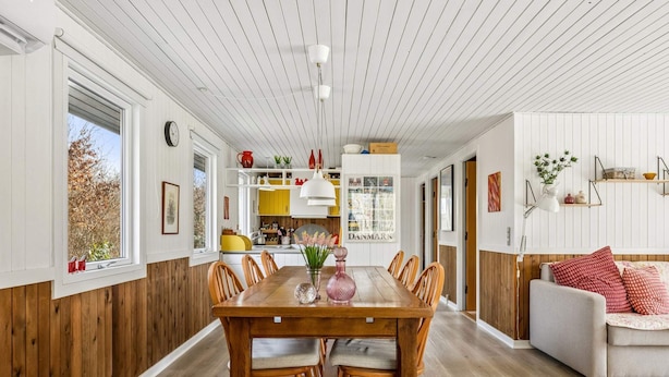Un comedor con una mesa y sillas de madera, una cocina al fondo y una sala de estar con sofá y cojines.