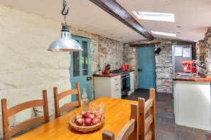 A rustic kitchen with stone walls, a wooden dining table with chairs, a hanging light fixture, and a small island with a sink. - Y Cwtch (Dolgellau)