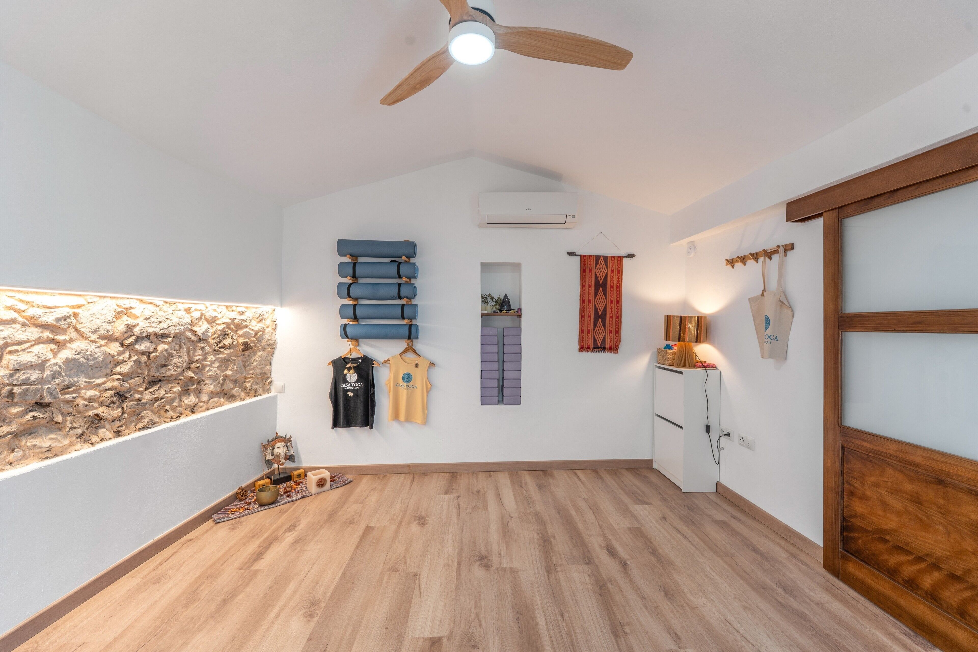 Une pièce avec un mur en pierre, un plancher en bois, un ventilateur de plafond, une armoire blanche et des serviettes suspendues.