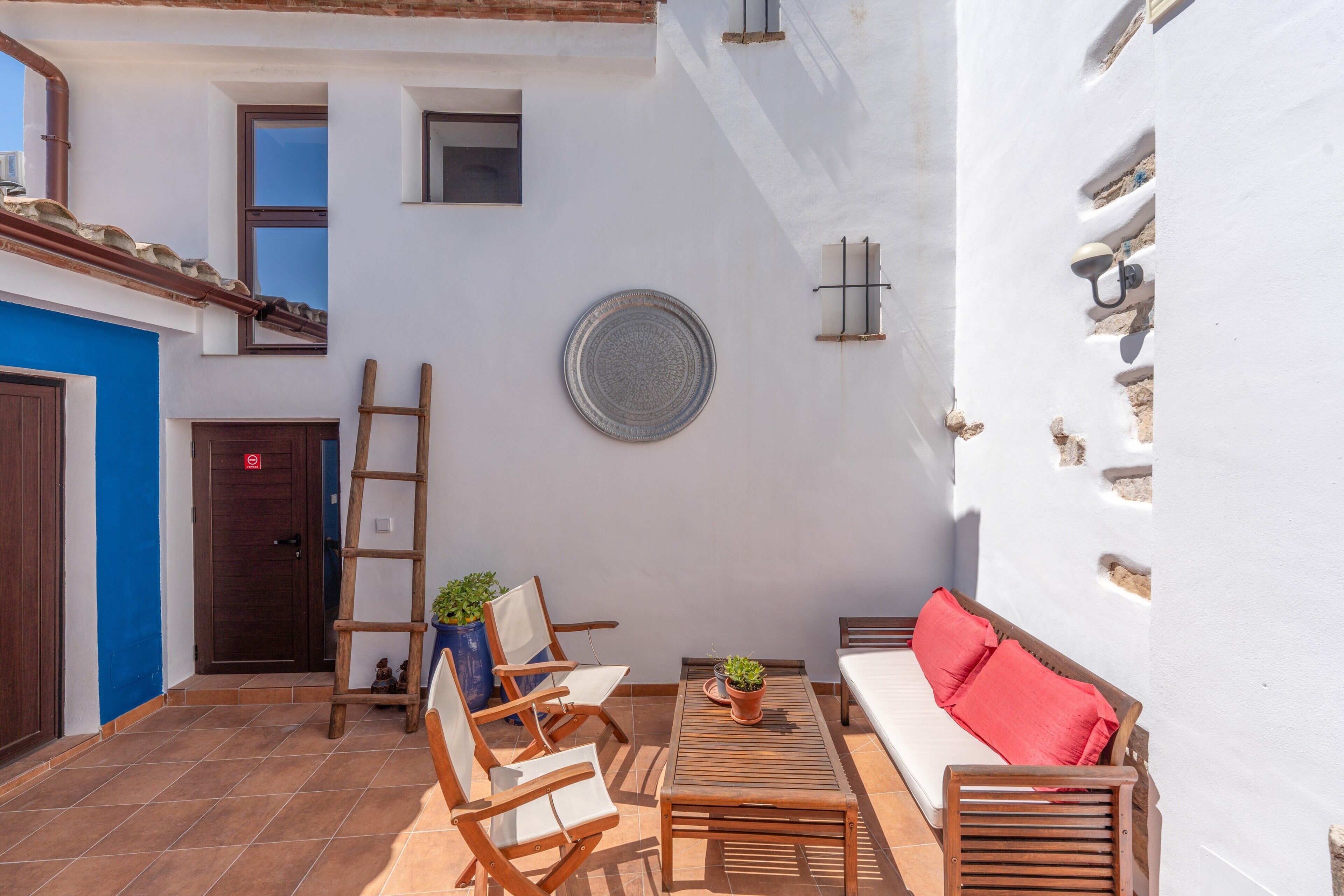 A courtyard with a wooden bench, a small table with a potted plant, a sofa with red cushions, a wooden ladder, and a wall-mounted decorative plate.