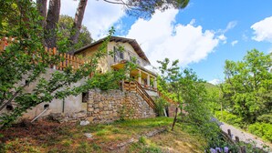 Ein Steinhaus mit einer Holztreppe, die zu einem Balkon führt, umgeben von Grünpflanzen und Bäumen.
