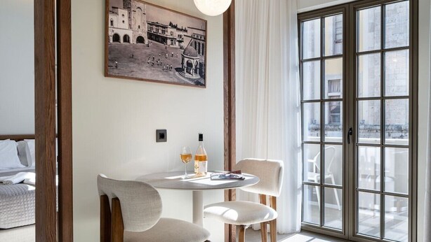 A hotel room with a bed, a round table with two chairs, a bottle of wine, and a painting of a historical building. - Volaka Luxury Suites `Ruby` (Kos)