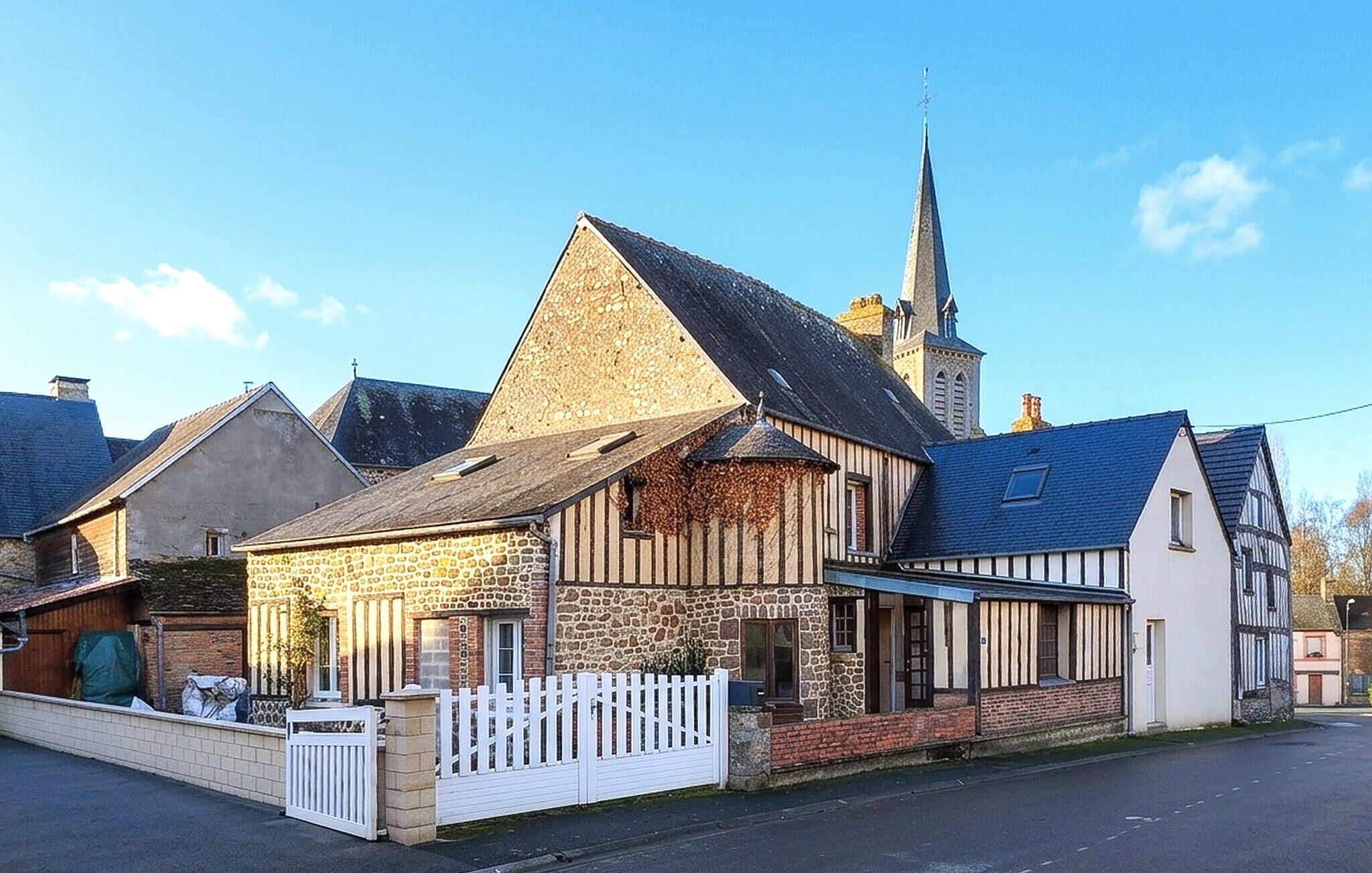 A traditional stone building with a steeply pitched roof, a white picket fence, and a church steeple in the background.