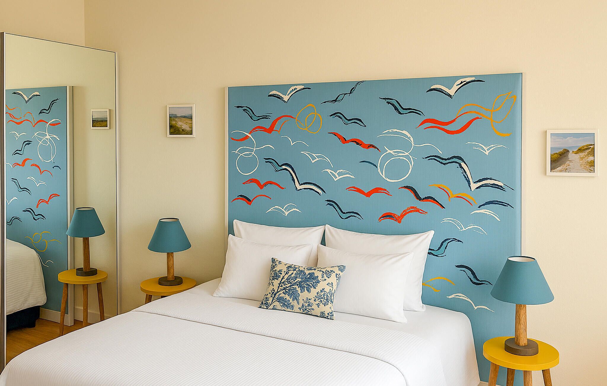A hotel room with a large bed, two bedside tables with lamps, a headboard with a bird pattern, and two framed pictures on the wall.