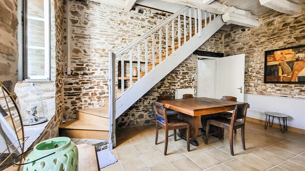 A rustic room with a stone wall, a wooden staircase, a dining table with chairs, and a small stool. - Charmante maison au calme (Saint-Yrieix-la-Perche)
