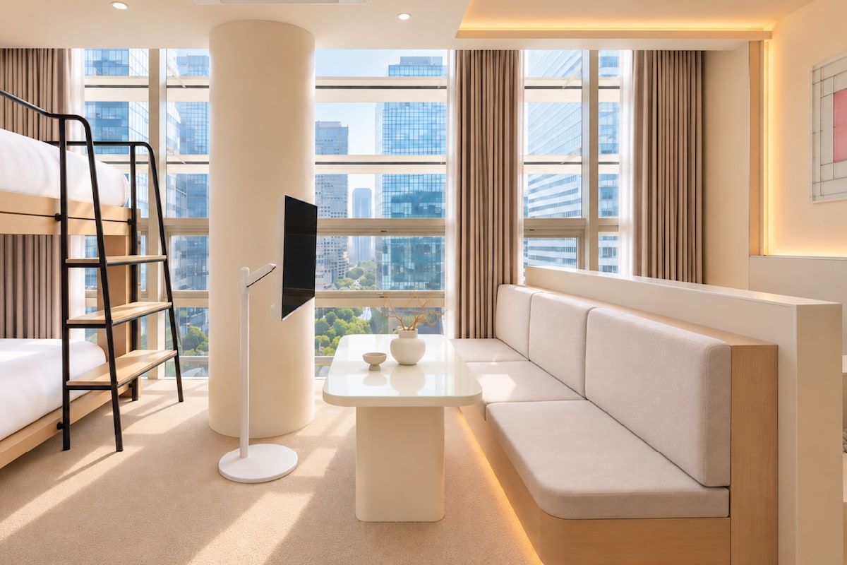 Hotelkamer met een stapelbed, een flatscreen televisie, een bank, een tafel en een groot raam met uitzicht op de stad.