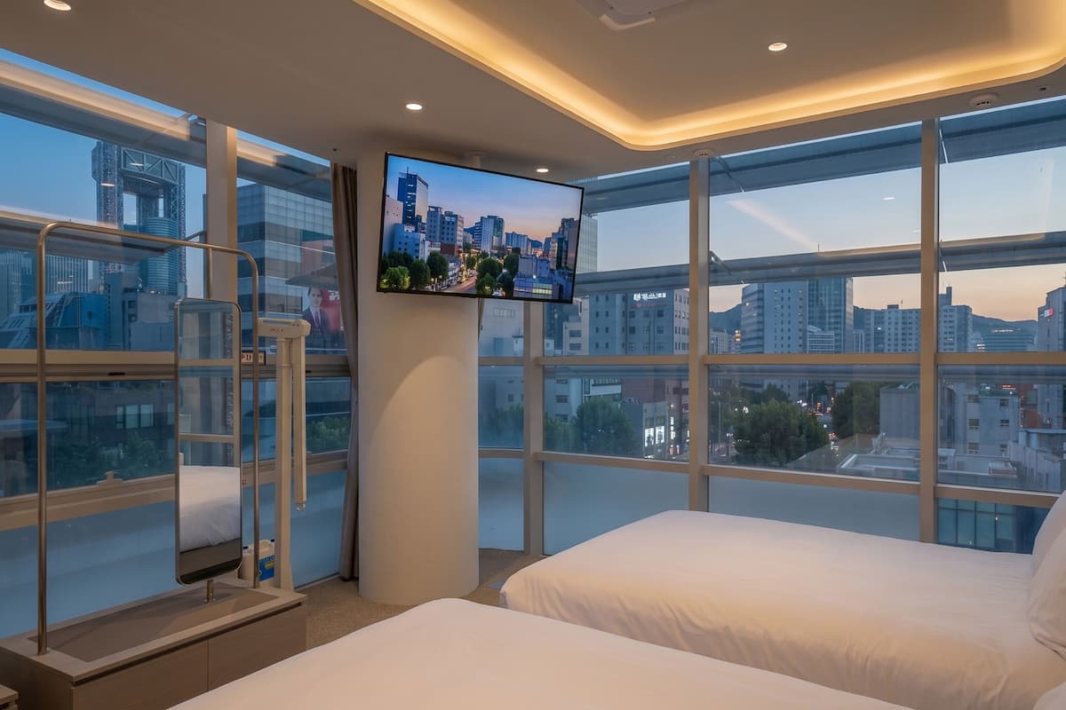 Habitación de hotel con un ventanal grande, una cama, un televisor y un balcón con barandilla.