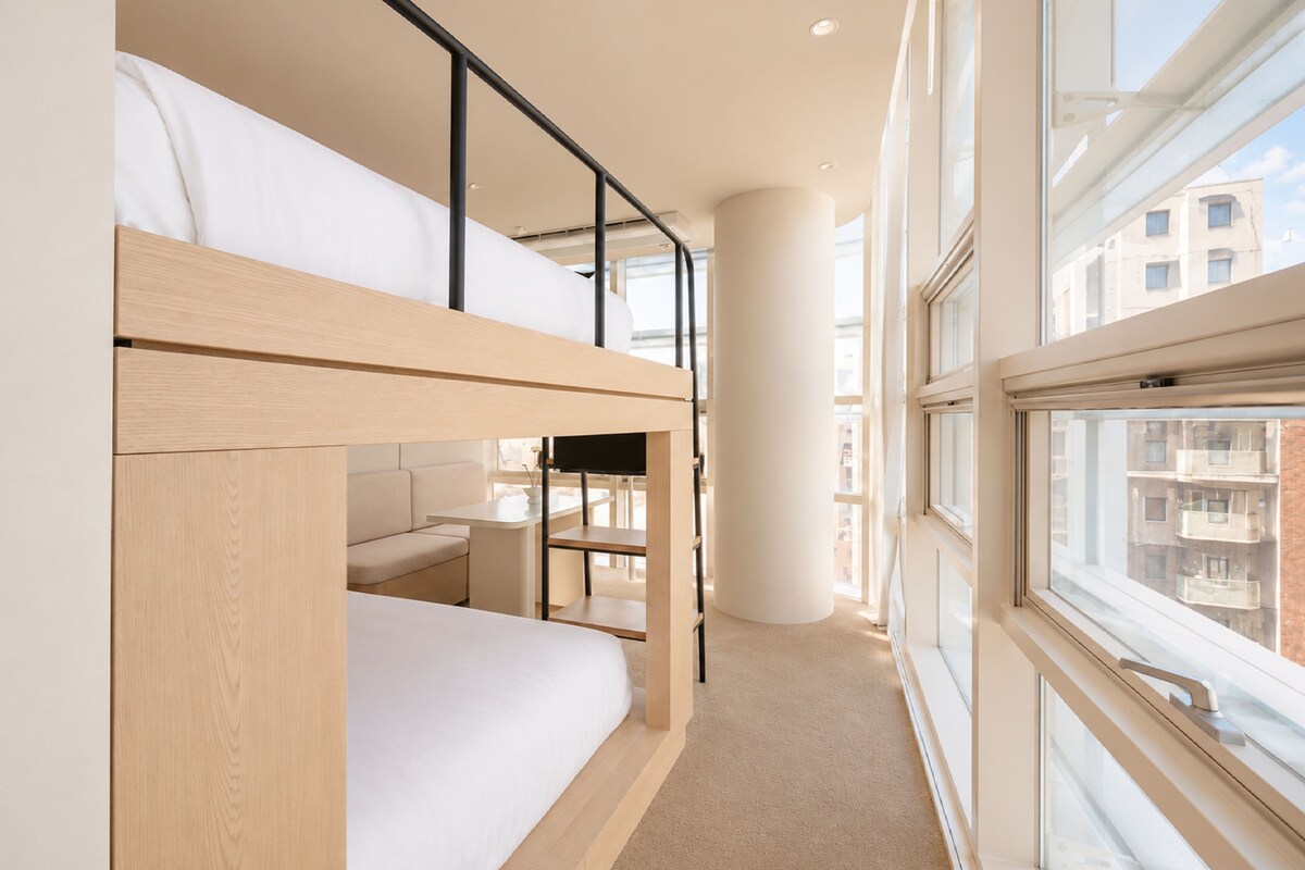 Een modern interieur met een stapelbed, een bureau en een groot raam met uitzicht op gebouwen.