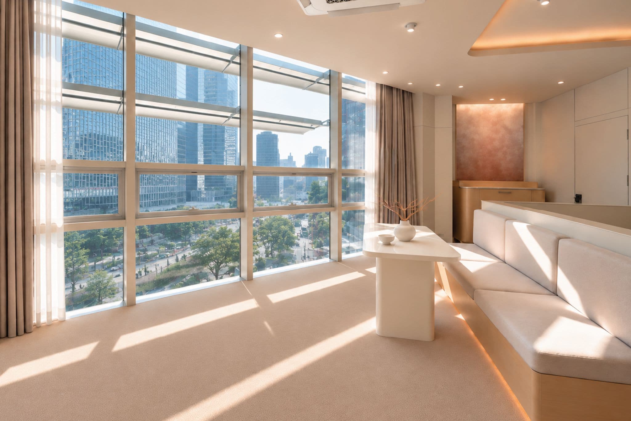 A modern hotel room with large windows, a sofa, a small table, and a view of the cityscape.