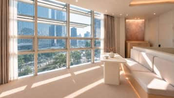 A modern hotel room with large windows, a sofa, a small table, and a view of the cityscape.