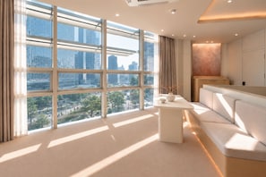 Quarto de hotel moderno com grandes janelas, um sofá, uma mesa pequena e vista para a paisagem urbana.