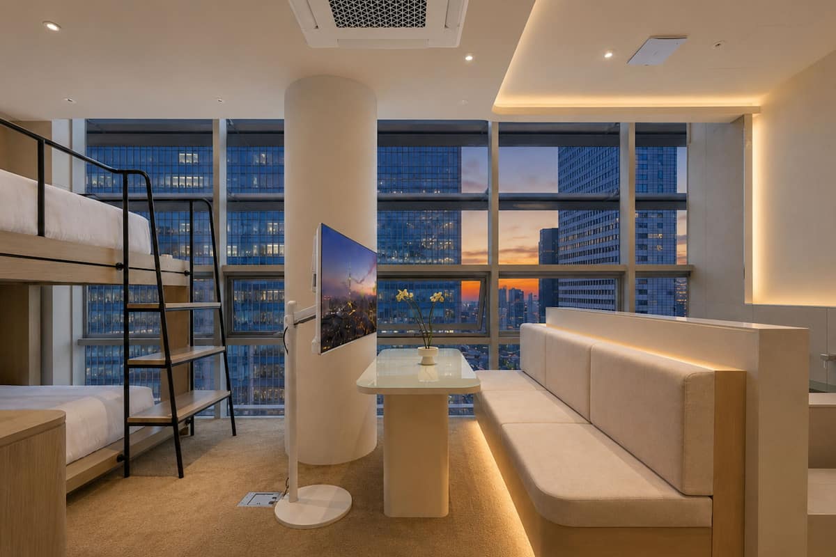 Una habitación de hotel moderna con una cama litera, un escritorio, un sofá y ventanas amplias con vistas a la ciudad.