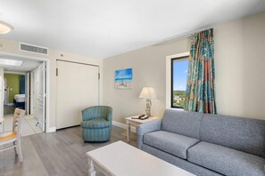 Ein Zimmer mit grauem Sofa, blauem Sessel, kleinem Tisch mit Lampe, einem Bild an der Wand und einem Fenster mit Vorhängen.