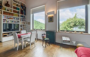 Ein Zimmer mit Bücherregal, Esstisch mit Stühlen, Heizkörper und großen Fenstern mit Blick auf Bäume.
